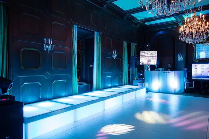 A modern event space with a lit up runway stage, elegant chandeliers, dark paneled walls, green curtains, and a DJ booth with screens displaying graphics, all bathed in blue lighting.