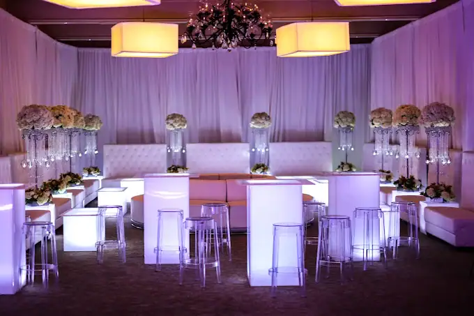 A modern event lounge with white draped walls, glowing illuminated tables, clear acrylic stools, white couches, floral arrangements in tall vases, and soft purple lighting under elegant chandeliers.