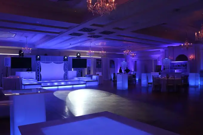 A large, elegant banquet hall is lit with purple and blue lights. The room features chandeliers, a dance floor, a stage with screens, and tables with chairs arranged around the perimeter.