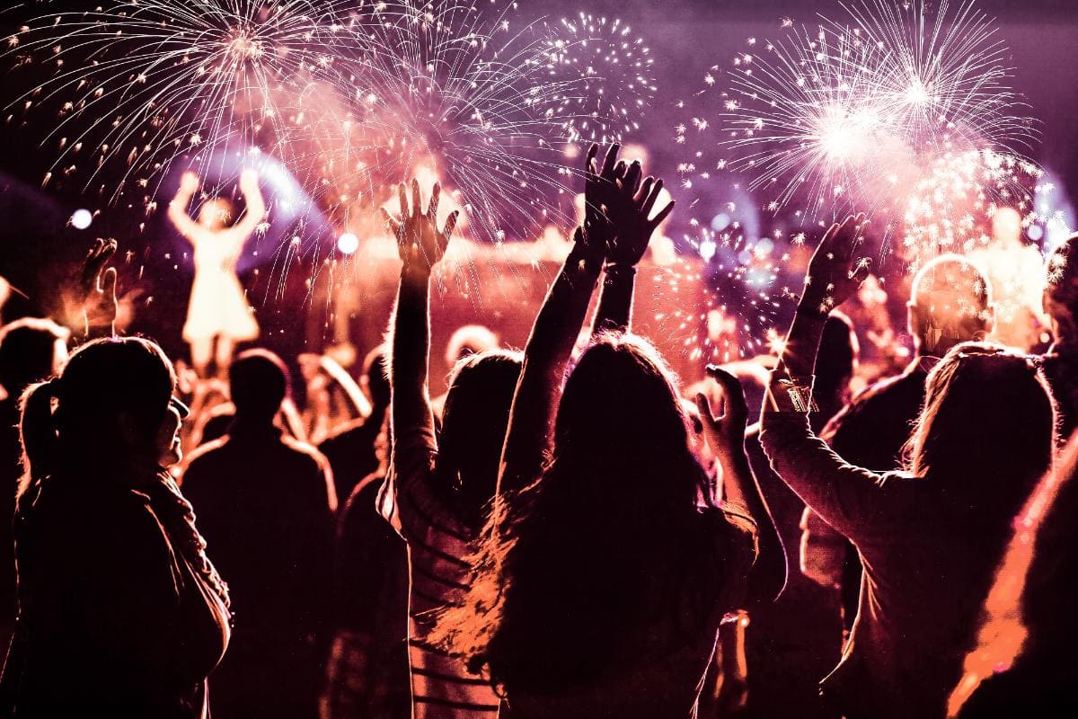 A crowd of people with raised arms celebrate at night, illuminated by colorful fireworks bursting in the sky above a stage with a performer in the background.