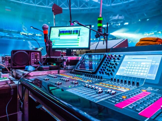 An audio mixing console with colorful buttons and screens, microphones, speakers, and computer monitors set up in a large stadium, illuminated by vibrant stage lights.