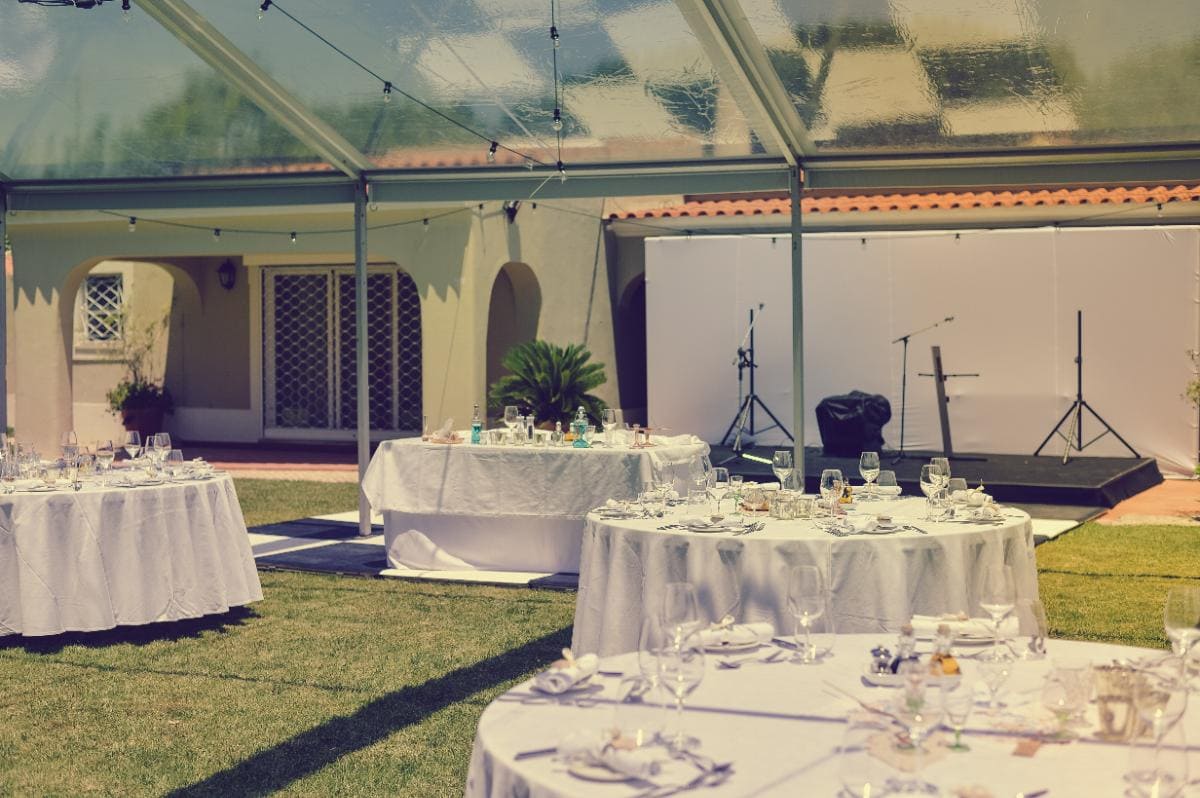 Outdoor event setup under a clear tent, with round tables covered in white cloths, place settings, and glassware. A small stage with microphones stands near a cream colored building in the background.