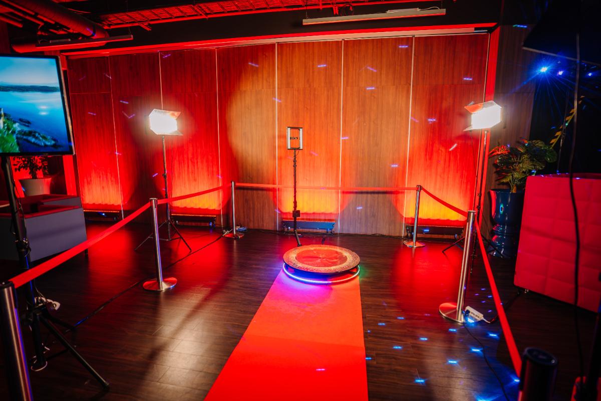 A red carpet leads to a round platform surrounded by red ropes, with bright lights and a camera setup in a modern room with red and blue ambient lighting.