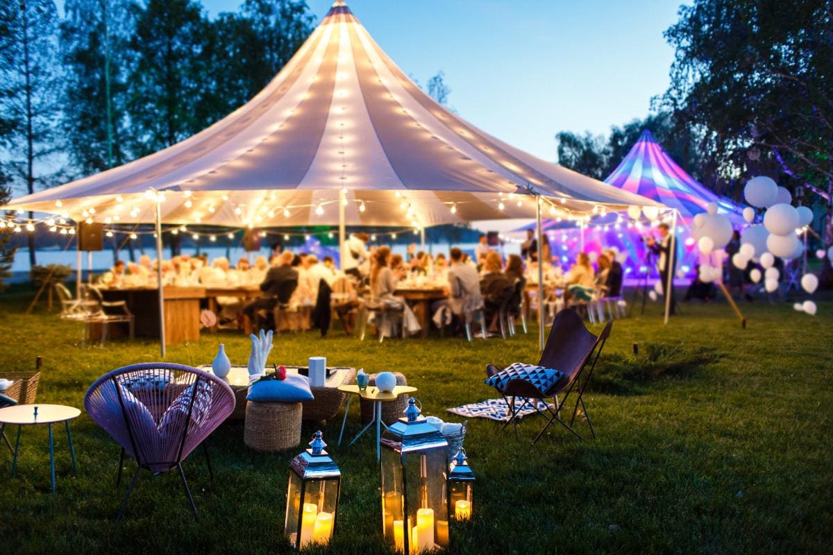 A festive outdoor event at dusk with people dining under a large white tent decorated with string lights. Lanterns, chairs, and cushions are arranged on the grass, and trees surround the area.