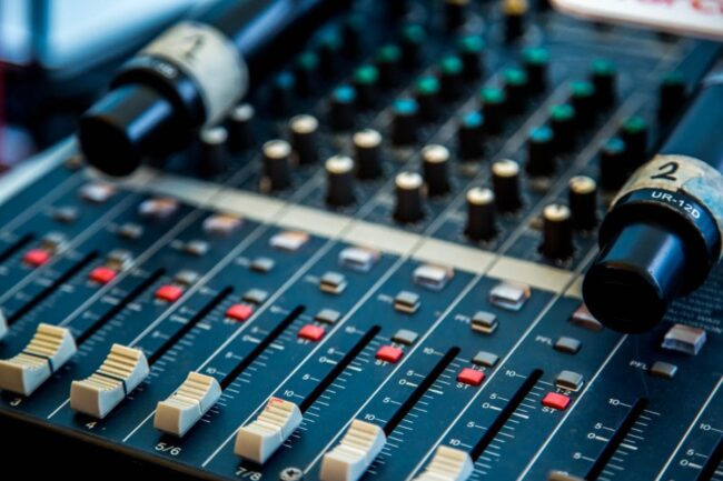 Close up of an audio mixing console with sliders, knobs, and buttons. Two microphones labeled "1" and "2" rest on top, partially covering the panel. The scene suggests a live or studio sound setting.