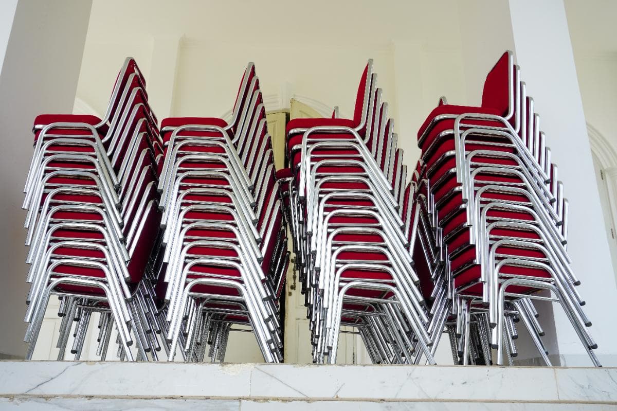 Stacks of red cushioned chairs with silver metal frames are arranged in several tall piles against a white wall on a white tiled platform.