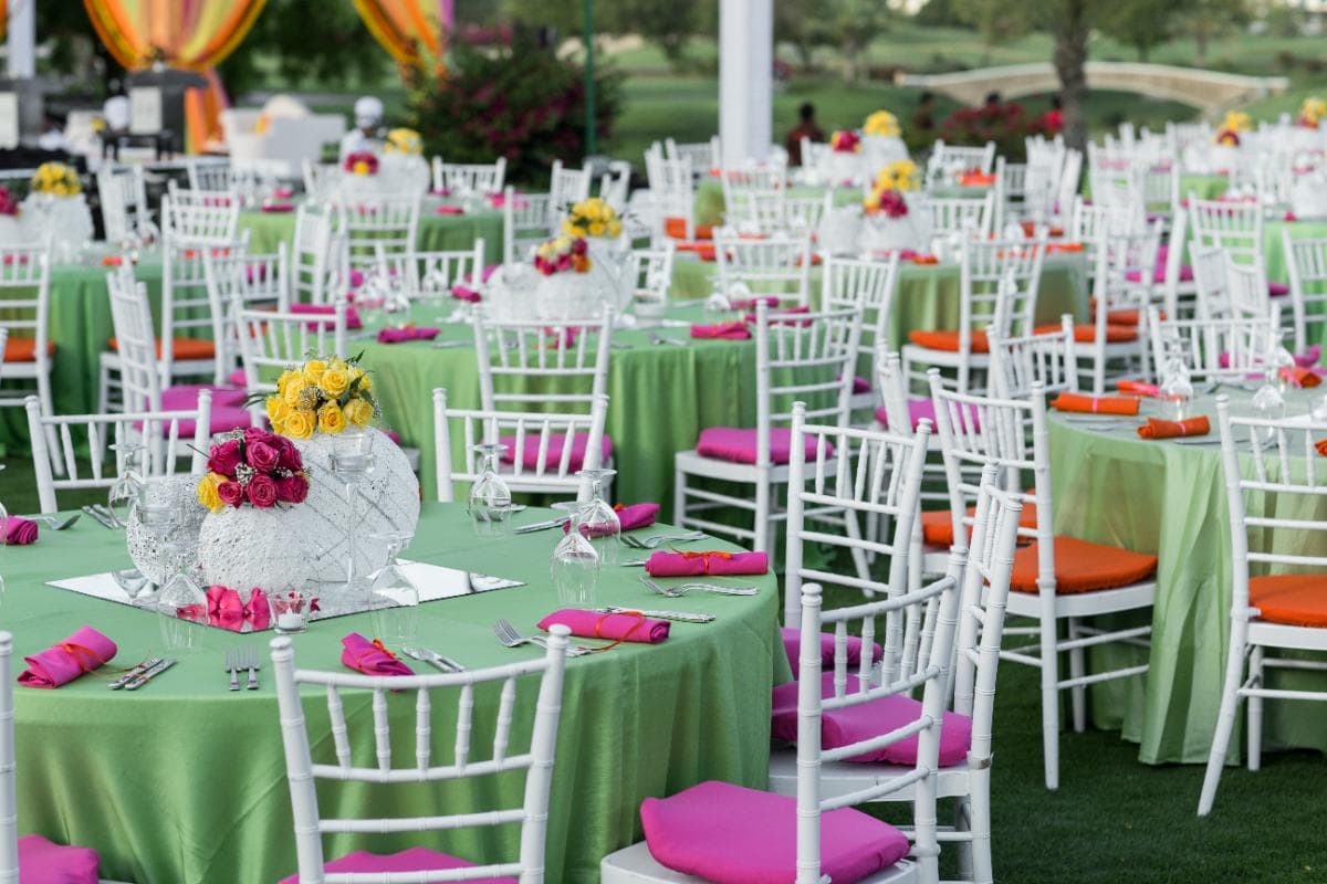 Outdoor event setup with round tables covered in green tablecloths, white chairs with orange seats, and pink napkins. Centerpieces feature bright flowers in yellow and pink tones. The scene is festive and colorful.