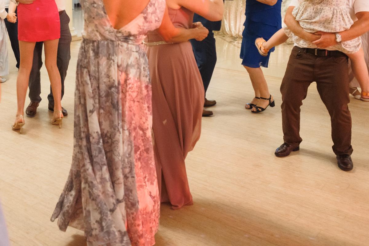 People dressed in semi formal attire are dancing together on a light colored floor. One person is holding a child, while others are paired or solo, enjoying the event. Only the lower halves of their bodies are visible.