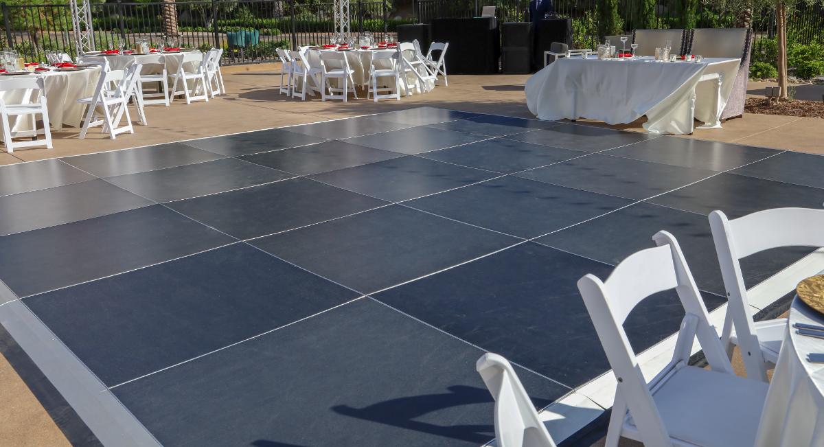 Outdoor event setup with a large empty dance floor surrounded by white chairs and tables with red accented place settings under sunny weather.