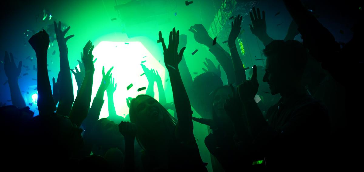 A crowd of people dance with raised hands in a dark club, illuminated by green and blue lights, with confetti falling around them.