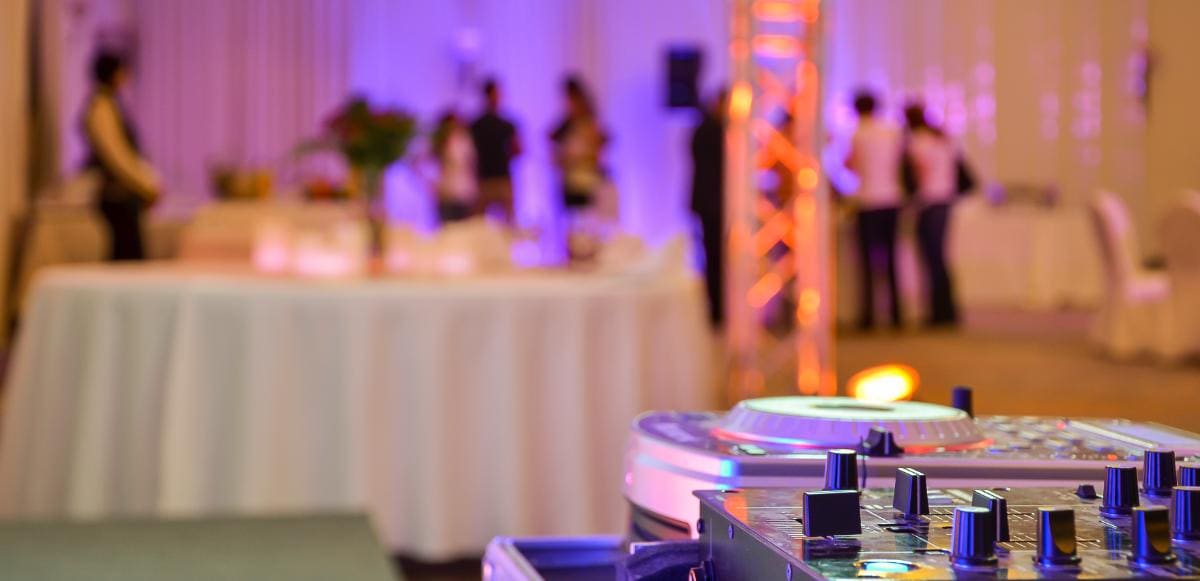 A DJ mixer sits in the foreground at an event venue with round tables and people mingling in the softly lit, purple and orange toned background.