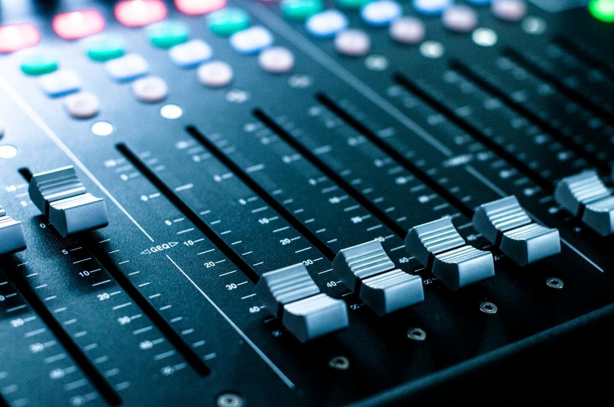 Close up of an audio mixing console showing several silver faders, buttons, and colorful lights, used for controlling sound levels in music or broadcast production.