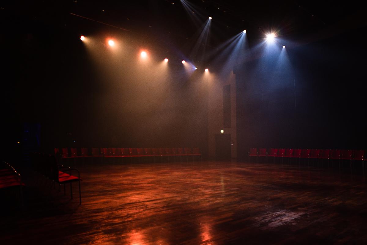 A dimly lit, empty theater with rows of red chairs along the walls and colorful stage lights illuminating the polished wooden floor in a smoky atmosphere.