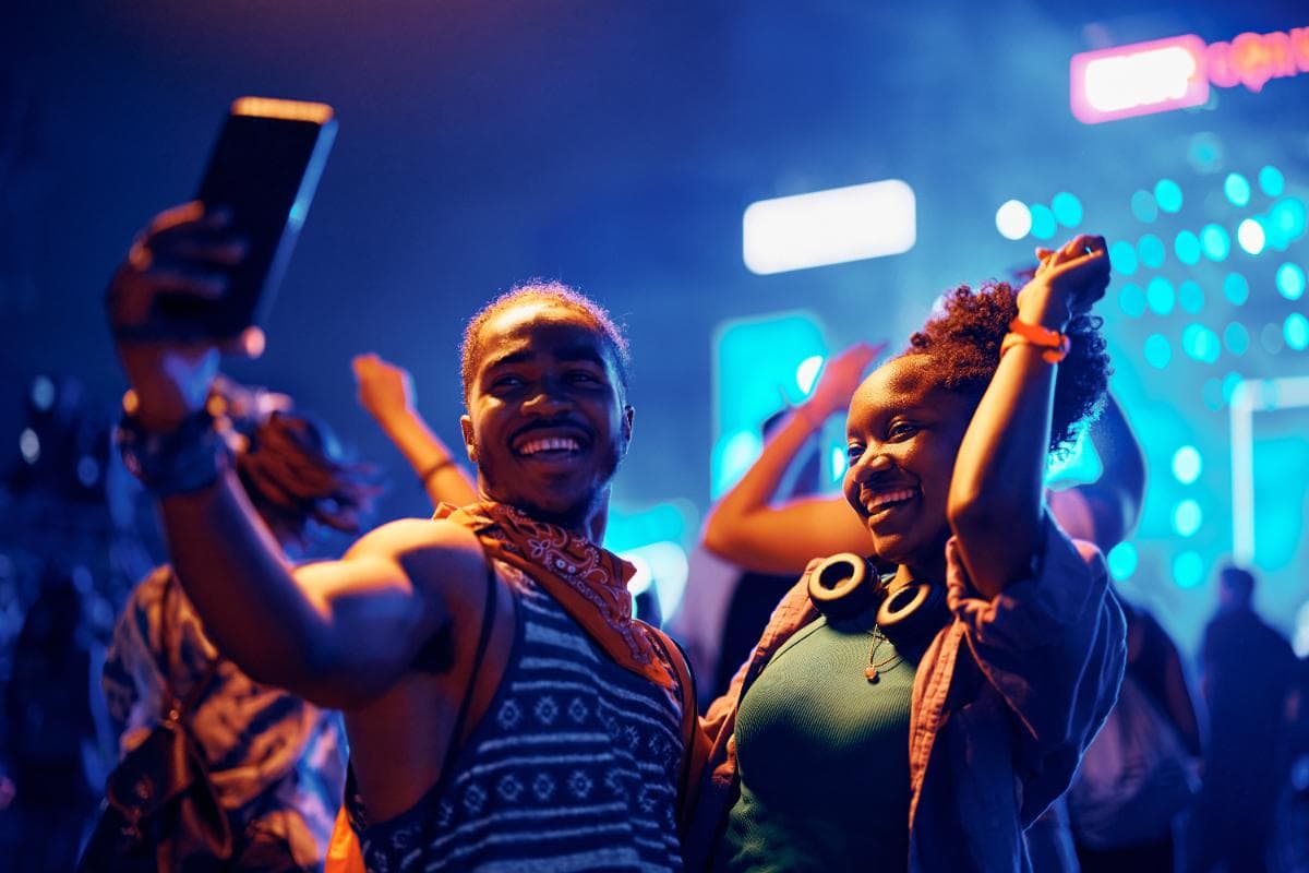 Two people smile and pose for a selfie at a lively, colorful event or concert, with bright neon lights and a crowd in the background. They appear joyful and energetic, enjoying the festive atmosphere.