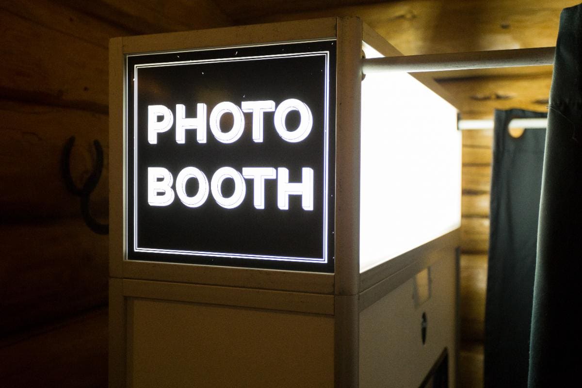 A photo booth with a bright, illuminated sign that reads "PHOTO BOOTH" in white letters, set against a wooden wall interior.