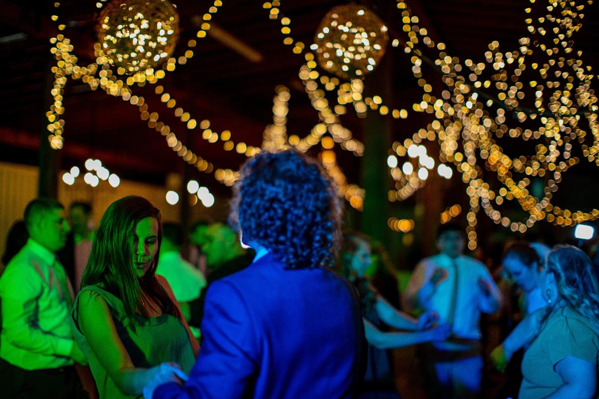 People dance under warm, glowing string lights at a crowded indoor event. The atmosphere is festive, with colorful lighting reflecting off the dancers and decorative hanging orbs above.