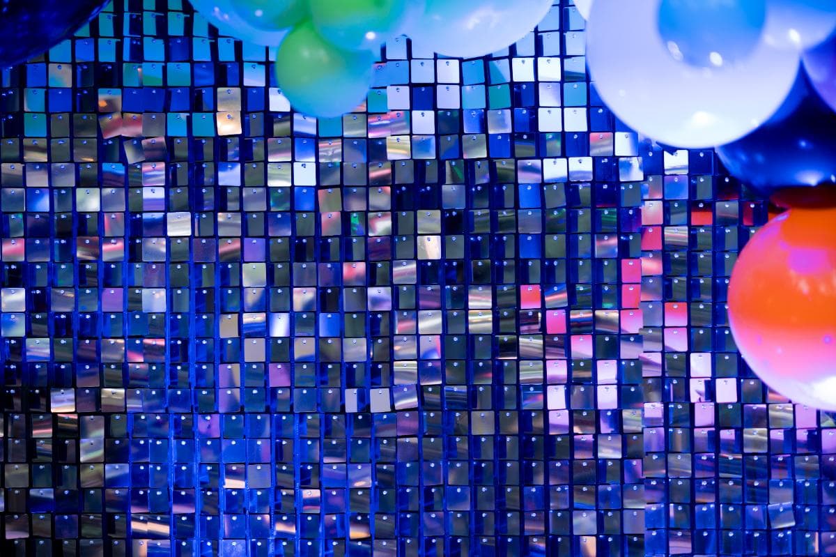 Colorful balloons hang at the top and right edges in front of a shimmering, reflective blue sequin backdrop, creating a festive and vibrant atmosphere.