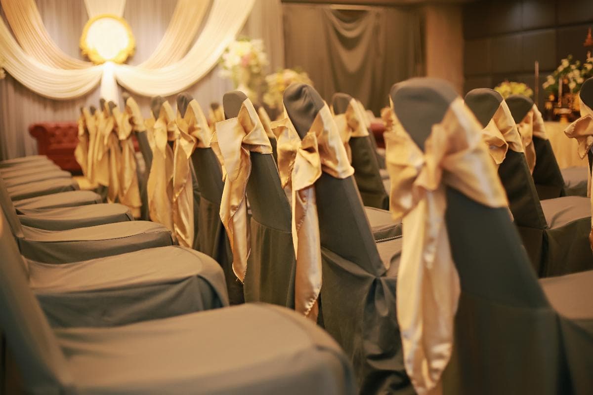 Rows of chairs covered in dark fabric with gold bows tied on the backs, arranged in a decorated indoor venue with soft lighting, draped curtains, and floral arrangements, suggesting a formal event or ceremony.