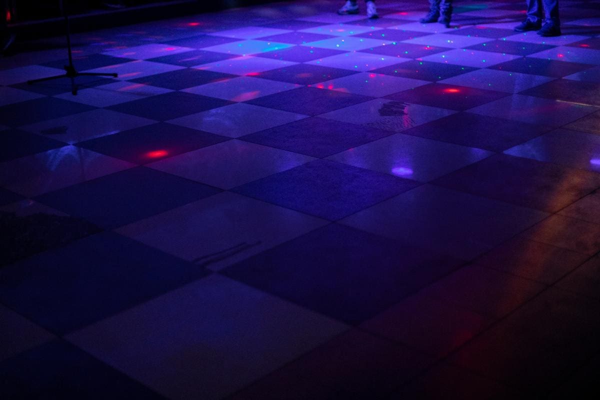 A dimly lit checkered dance floor with colorful disco lights reflecting on the tiles. The lower legs and feet of a few people are visible in the background.