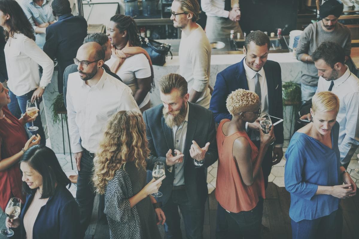 A diverse group of people in business attire mingle and converse at a social event. Some hold drinks, others use digital devices. The setting appears to be a modern, busy indoor café or bar.