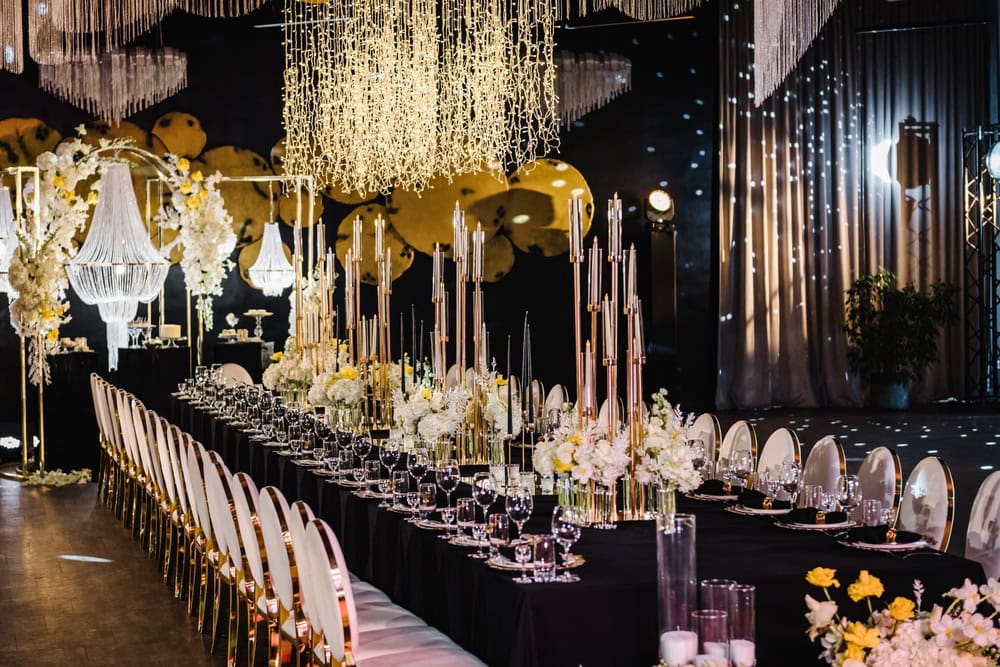 A long, elegant banquet table is set with white flowers, gold accents, glassware, and tall candles. Chandeliers and string lights hang above, creating a luxurious, festive atmosphere in a dark, ornate room.