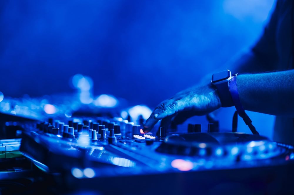 A DJ's hand adjusts controls on a turntable mixer, illuminated by blue stage lights, creating a lively and energetic atmosphere.
