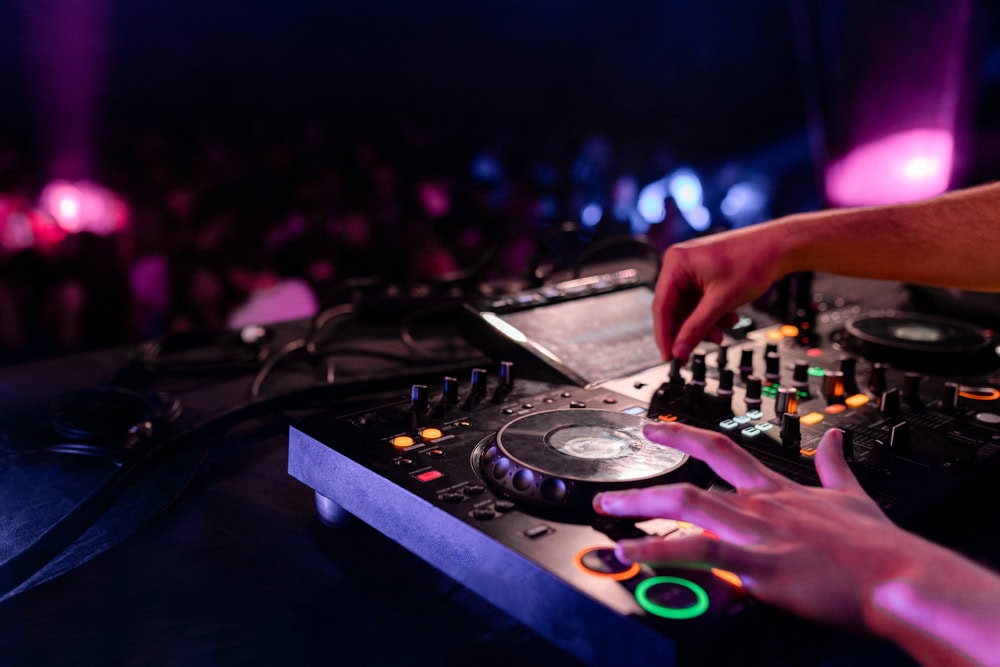 A DJ’s hands adjust knobs and buttons on a mixing console, with colorful lights illuminating the equipment and a blurred crowd in the background at a lively music event.
