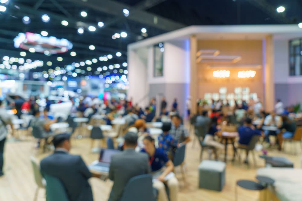 A blurred image shows a busy indoor event with many people sitting at tables and walking around. Bright lights and a modern booth setup are visible in the background, creating a lively atmosphere.