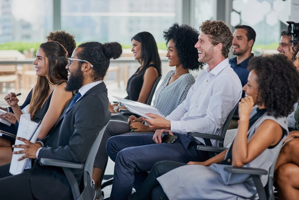 A group of diverse professionals sit in chairs, smiling and listening attentively during an indoor seminar or business meeting. Some hold notepads and pens, appearing engaged and relaxed in a bright, modern setting.