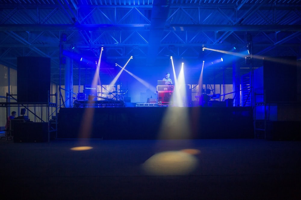 A DJ stands behind a turntable on a stage, illuminated by blue and purple spotlights, in an empty indoor venue with visible lighting equipment and metal beams.
