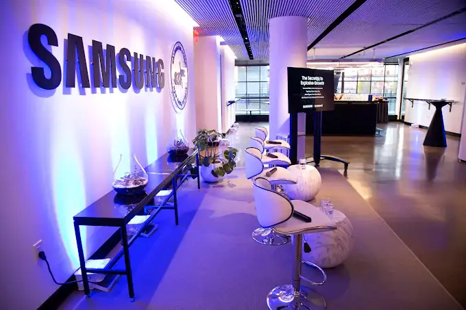 A modern, brightly lit event space with “SAMSUNG” on the wall, white chairs on marble bases, a black table with glass bowls, potted plants, and a large screen, with large windows in the background.
