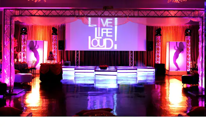 A vibrant event stage with purple lighting, glowing dance floor, and a large screen displaying "LIVE LIFE LOUD." Silhouettes of dancers appear on lit panels at each side, creating an energetic party atmosphere.
