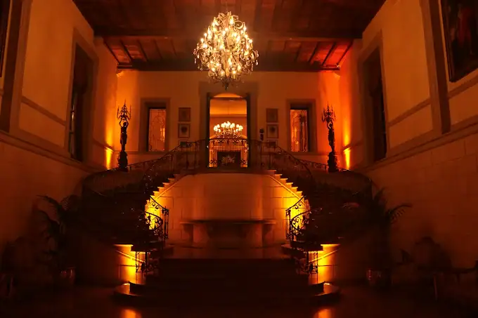 A grand staircase splits into two curved flights, illuminated by warm, amber lighting. An ornate chandelier hangs above, and elegant decor and columns frame the luxurious, classical interior.