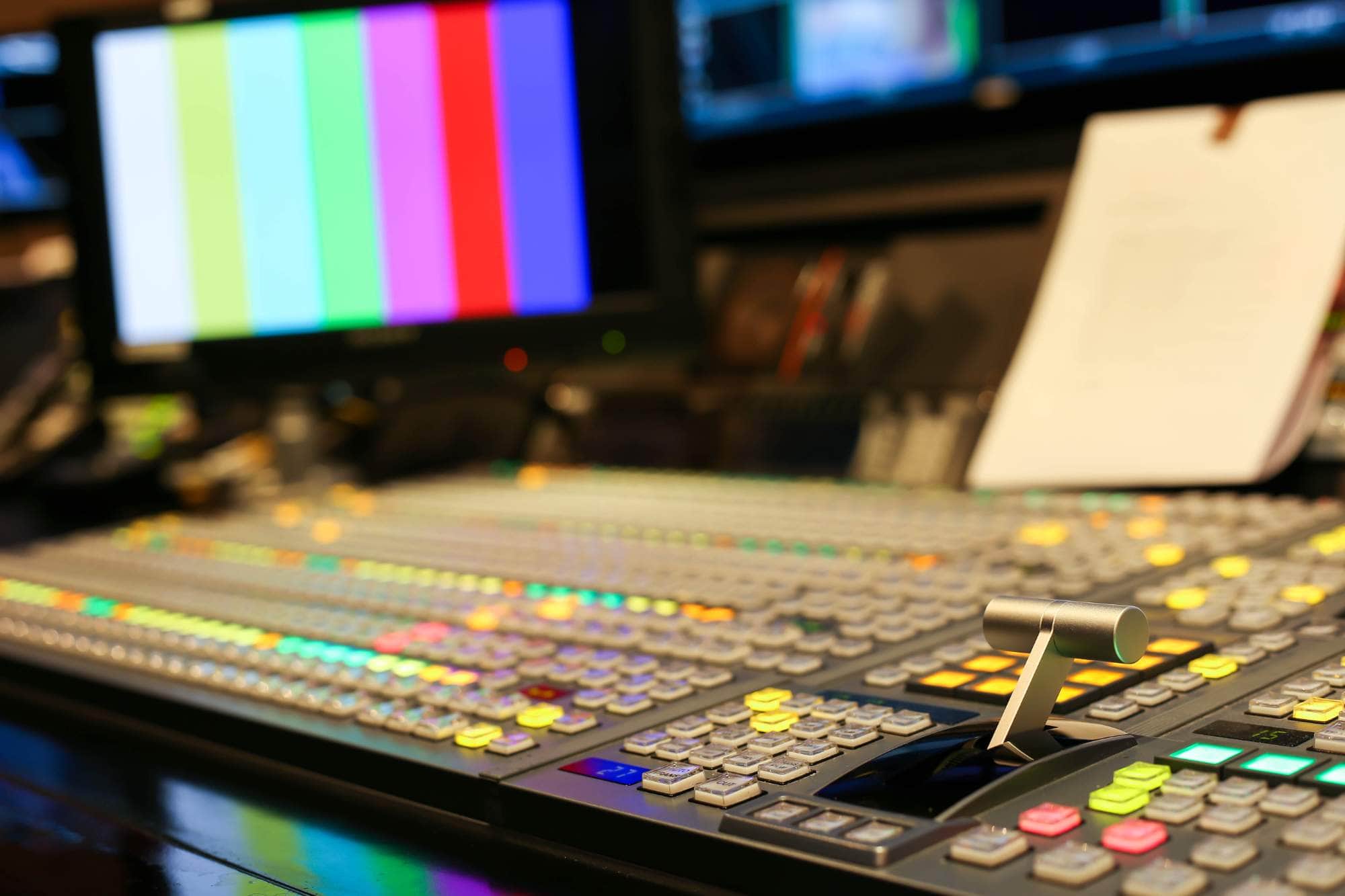 A close up of a television production control panel with colorful buttons and levers, and a monitor displaying vertical color bars in the background. A clipboard with paper is also visible.