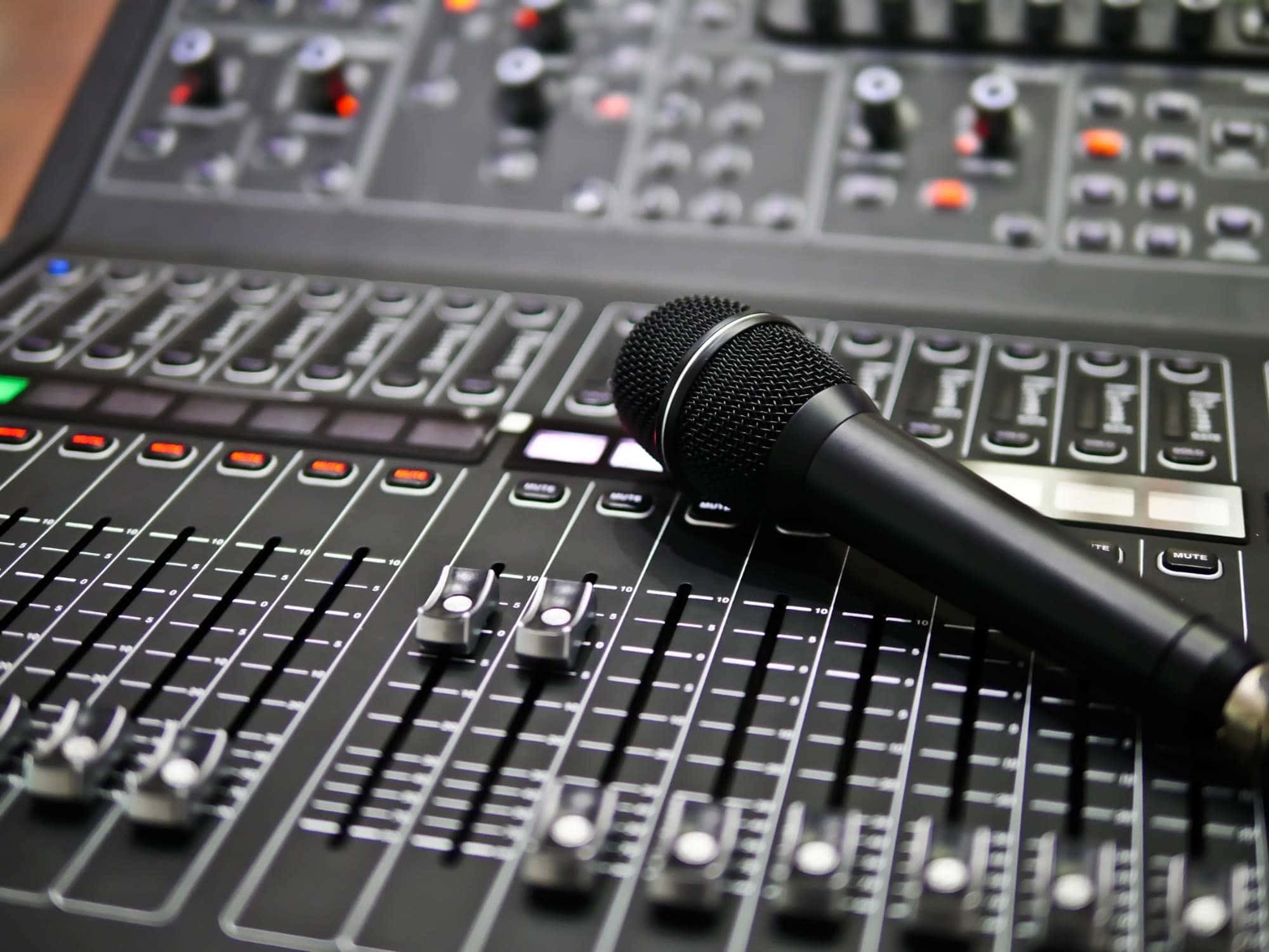 A black microphone resting on a sound mixing console with various sliders, buttons, and knobs, commonly used for audio production or live sound engineering.