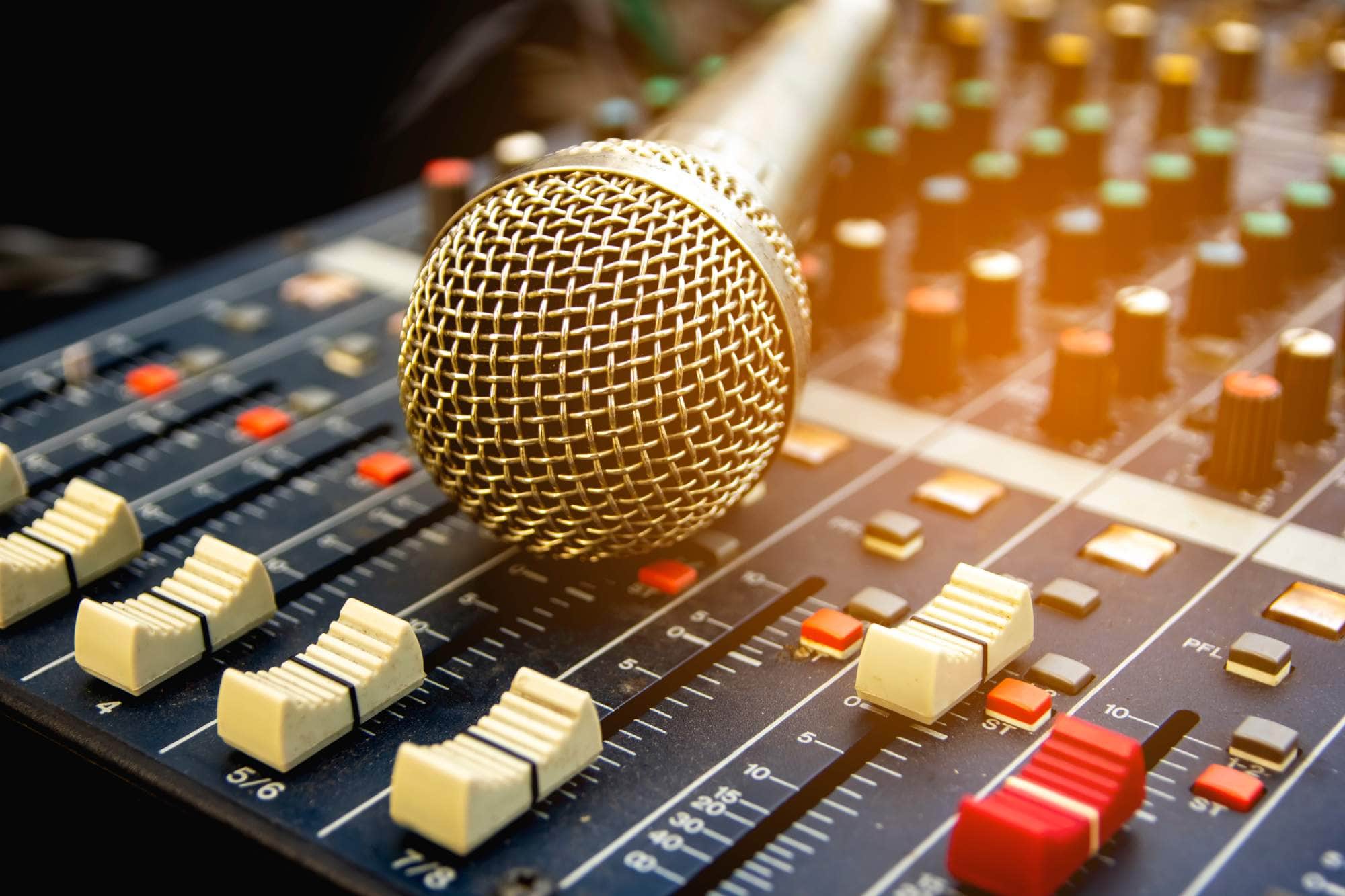 A close up of a microphone resting on an audio mixing console with various sliders, knobs, and buttons in different colors. Warm light is highlighting part of the scene.