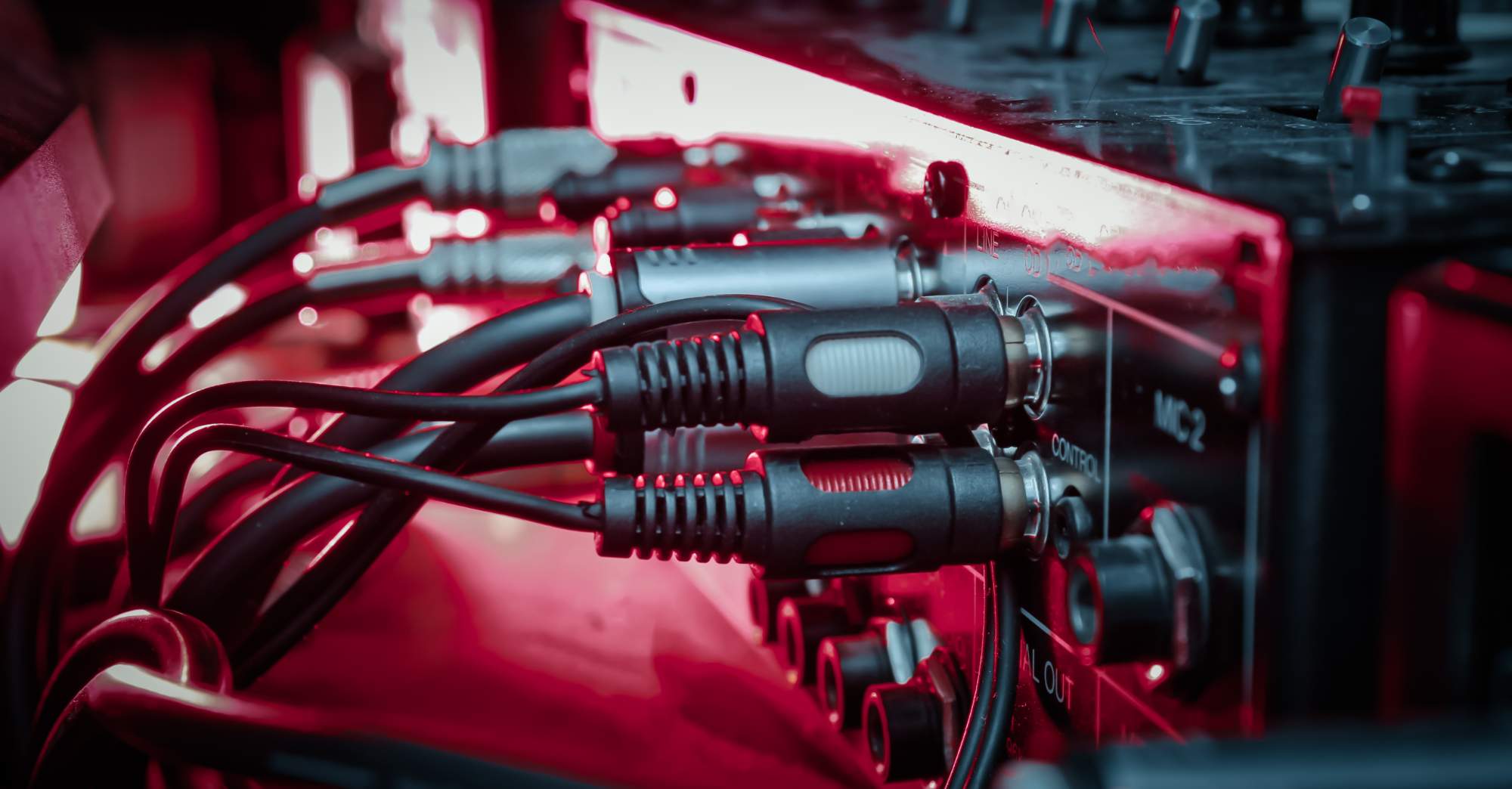 Close up of multiple audio cables plugged into a sound mixing console, with red and blue lighting reflecting off the equipment.