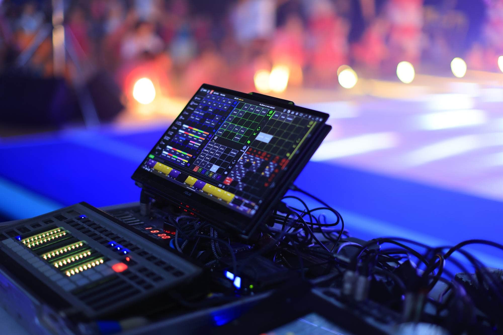 A close up of a lighting control panel and computer screen with colorful controls, set up at an event with a blurred, brightly lit stage and audience in the background.