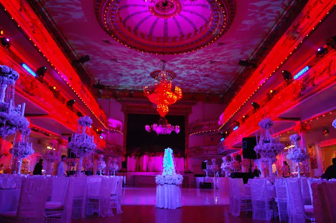 A grand ballroom decorated for an event, featuring elegant white tables and floral centerpieces, a large ornate chandelier, and dramatic pink and red lighting illuminating the ceiling and walls.