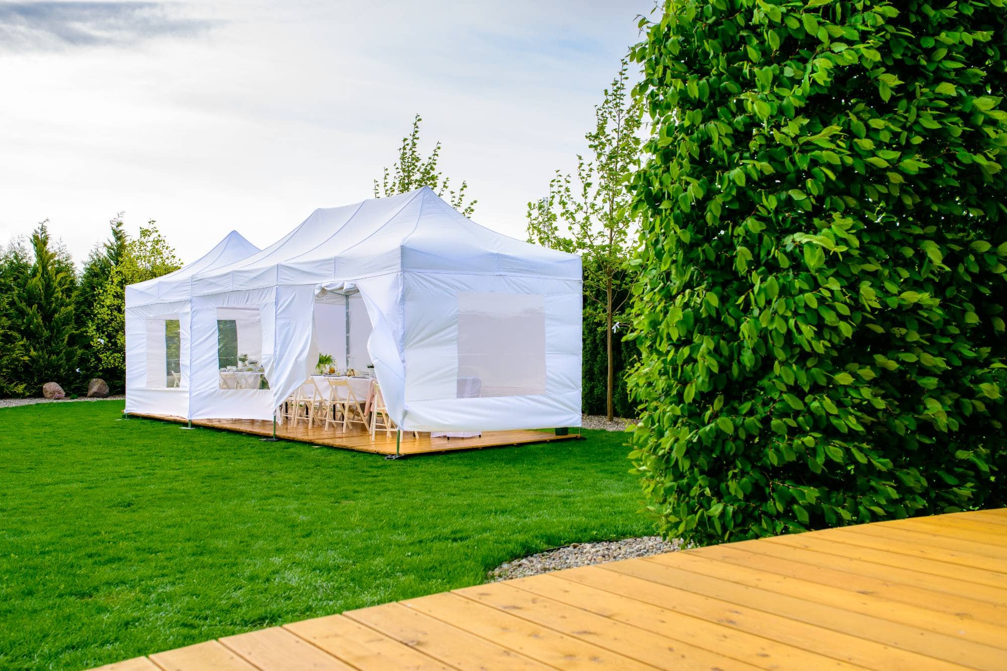 A large white event tent with clear windows stands on a lush green lawn. Inside, decorated tables and chairs are visible. Nearby, there is a wooden deck and tall leafy hedges surround the area.