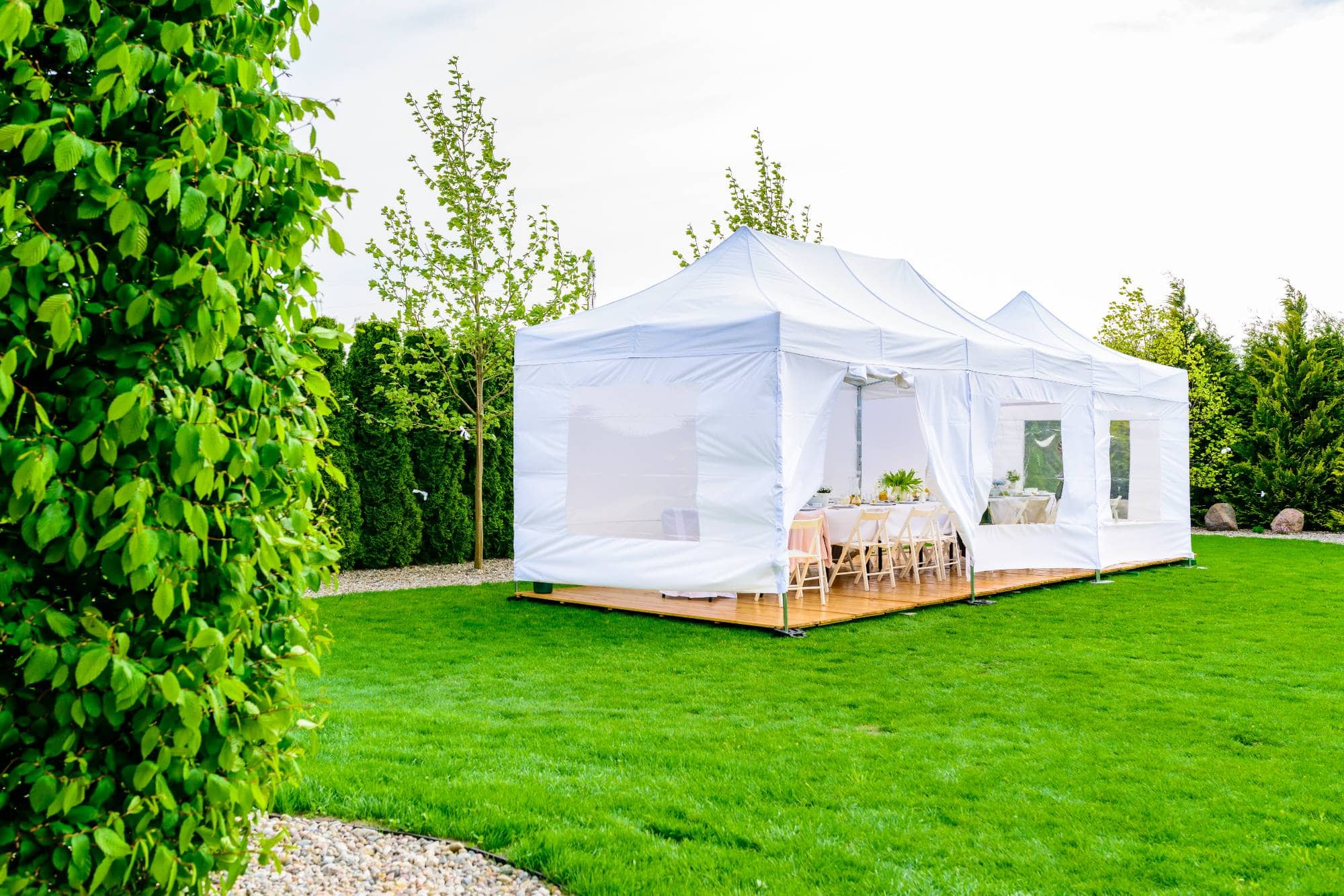 A white event tent with tables and chairs inside is set up on a green lawn, surrounded by trees and shrubs on a sunny day.
